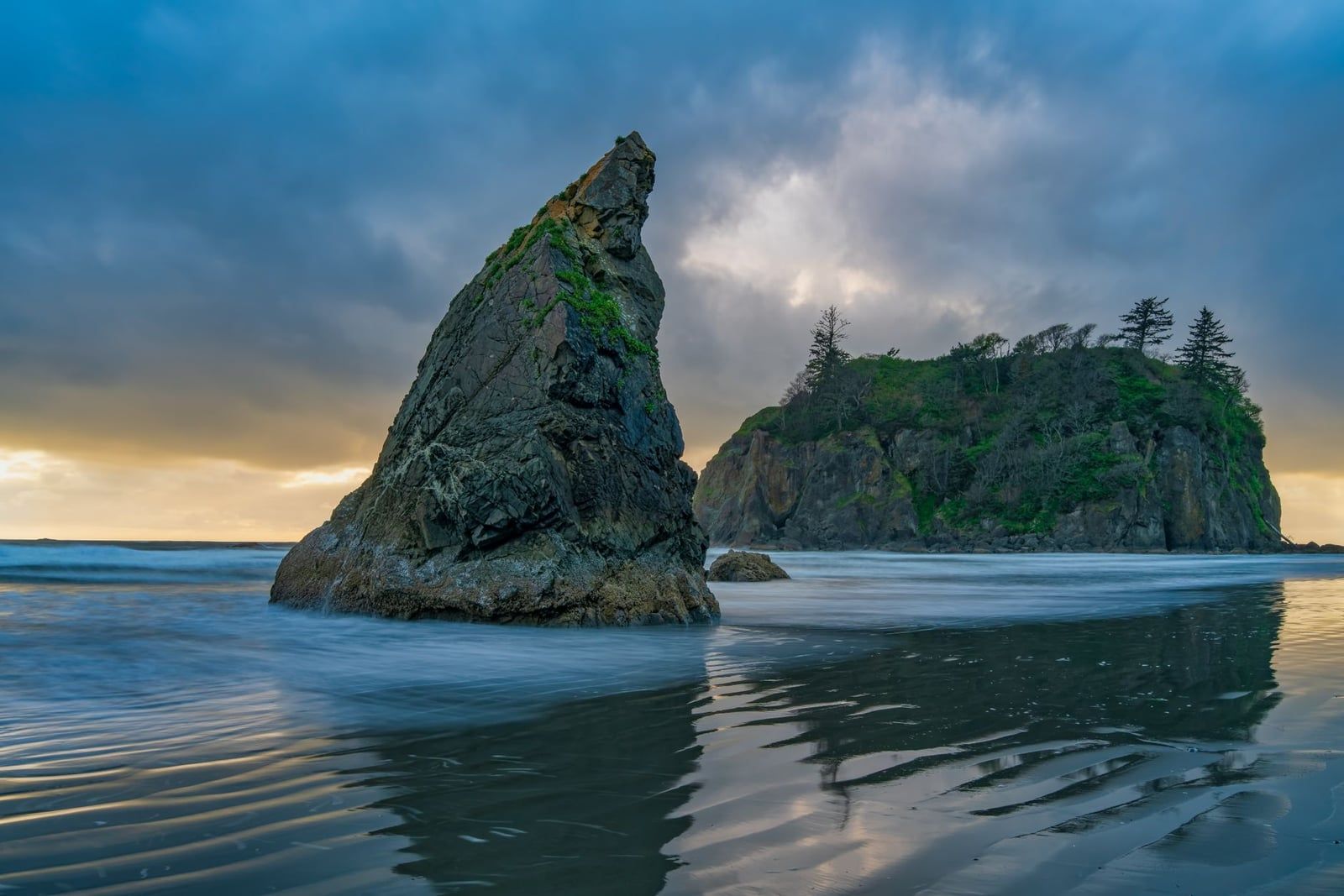 A jagged sea stack and forested island rise from the wet sand and incoming surf at Ruby Beach in Olympic National Park.