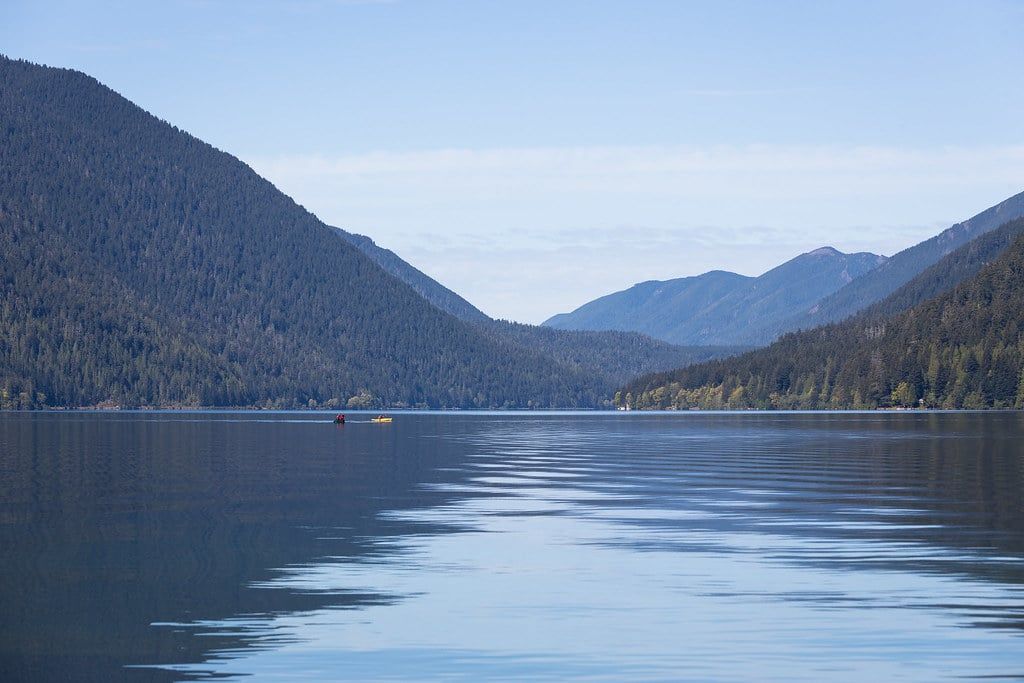 Two kayakers paddle across the calm blue waters of Lake Crescent surrounded by steep forested mountains in Olympic National Park.