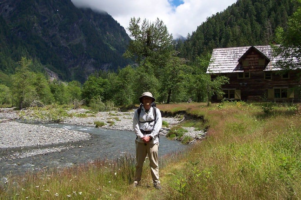 A backpacker stands near a riverbank before the historic wooden Enchanted Valley Chalet in Olympic National Park.