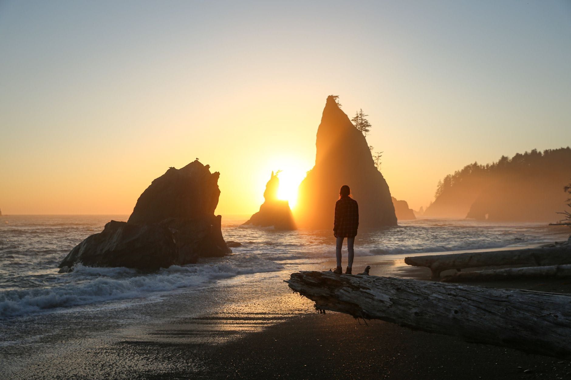 A person stands silhouetted on a driftwood log watching the sunset behind sea stacks along Rialto Beach in Olympic National Park.