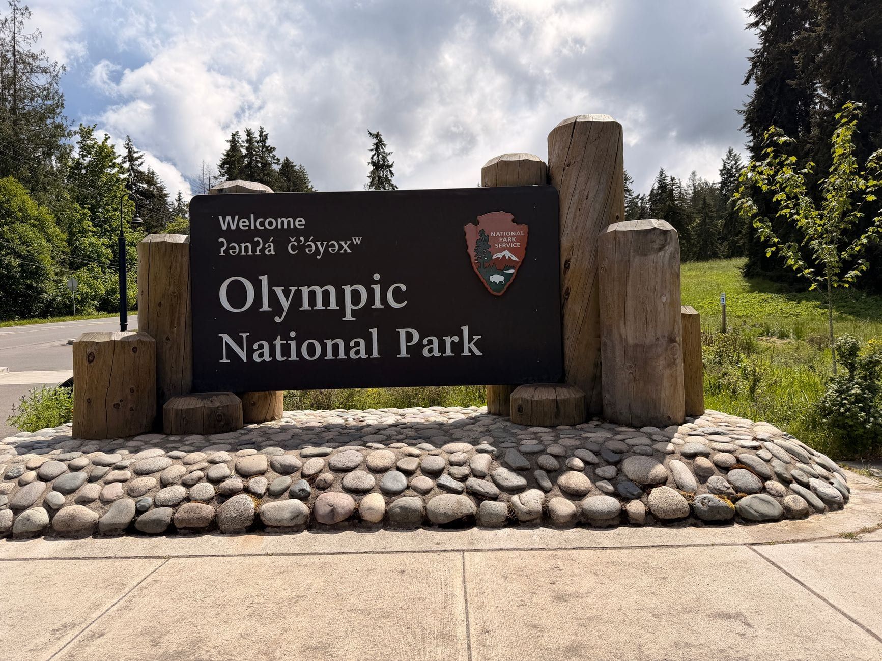 An official Olympic National Park entrance sign flanked by wooden posts and smooth stones beneath a partly cloudy sky.