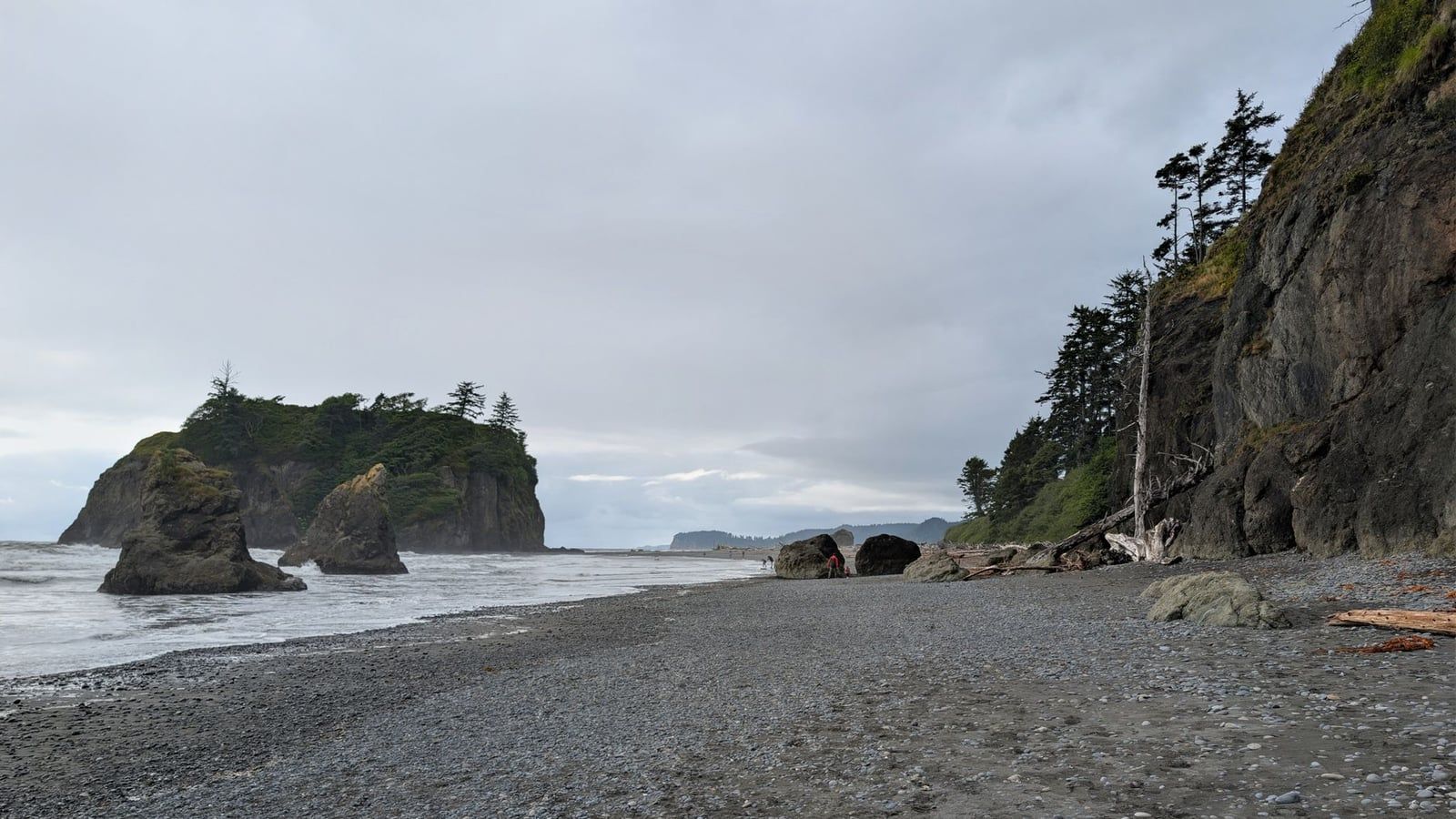 Sea stacks and a forested island rise from the ocean along the pebbled coastline of Rialto Beach in Olympic National Park.