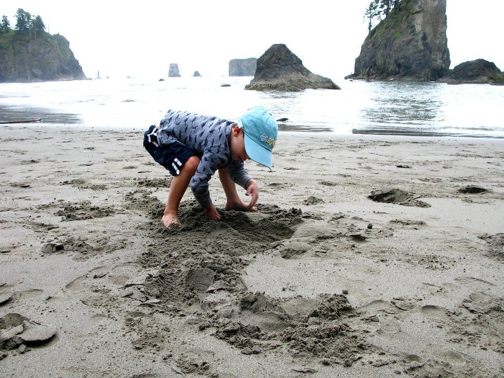 A young child plays in the sand on Ruby Beach with coastal sea stacks rising from the ocean in Olympic National Park.